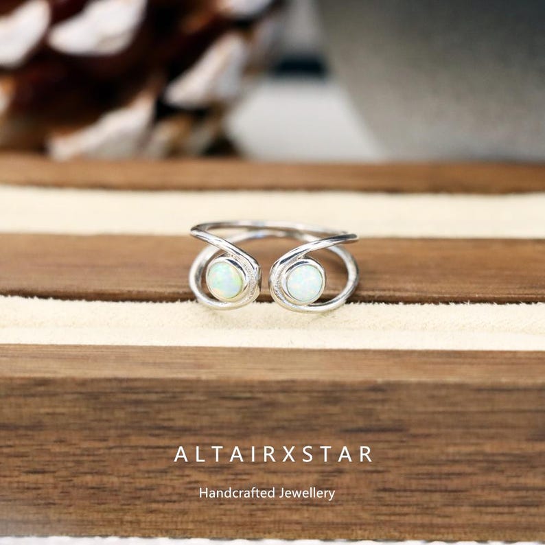 May include: A silver ring with two opal stones. The ring has a unique design with the stones set in open, swirling settings. The ring is displayed on a wooden surface with the text "ALTAIRXSTAR" and "Handcrafted Jewellery".