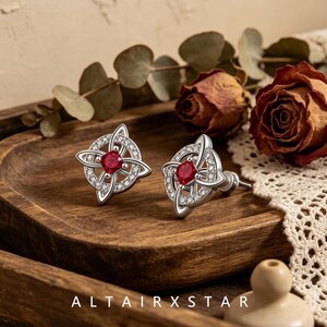 May include: A pair of silver stud earrings featuring a circular Celtic knot design encrusted with small clear stones and a central square-cut red gemstone. The text ALTAIRXSTAR is printed at the bottom of the image in white capital letters.
