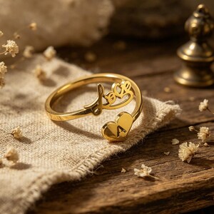 May include: A gold-colored ring with the word "Love" in a cursive font. The ring features a heart-shaped design with the letter "A" engraved on it. The ring is displayed on a beige cloth, with a wooden surface in the background.
