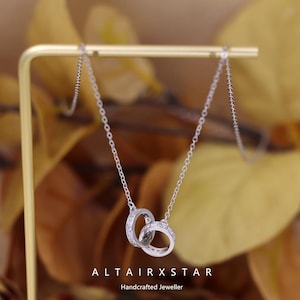May include: A silver necklace with two intertwined rings, each set with small, sparkling stones. The necklace is suspended from a fine chain, presented on a gold stand against a backdrop of blurred autumn leaves. The text "ALTAIRXSTAR Handcrafted Jeweller" is visible.