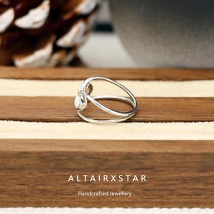 May include: A silver ring with a white gemstone, featuring a unique, open design. The ring has a double band that curves around the finger, with a small, oval-shaped gemstone set at the top. The ring is displayed on a wooden surface. The text "ALTAIRXSTAR Handcrafted Jewellery" is at the bottom.
