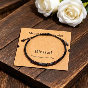 May include: A black Morse code bracelet with a "Blessed" card. The bracelet has a woven design with small, dark beads and reddish-brown accents. The card is brown with white text and is placed on a wooden surface.