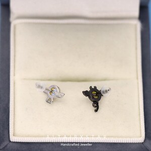 May include: A pair of stud earrings, one silver-toned and one black, each with a unique design. The silver earring has a stylized shape, while the black earring features a more intricate, curved design. The earrings are presented in a white jewelry box.