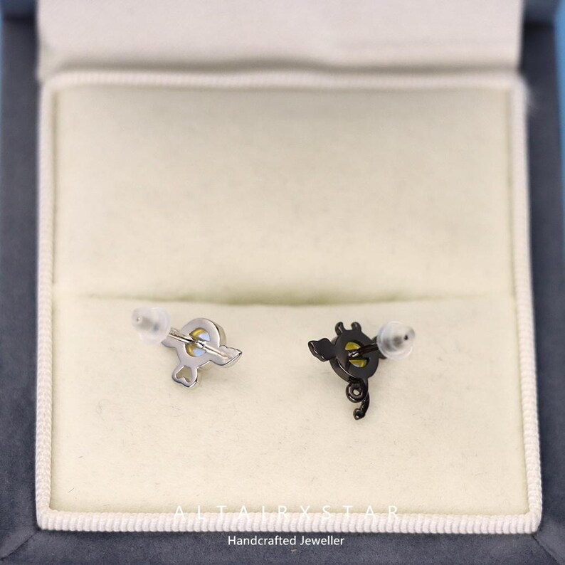 May include: A pair of stud earrings, one silver-toned and one black, each with a unique design. The silver earring has a stylized shape, while the black earring features a more intricate, curved design. The earrings are presented in a white jewelry box.