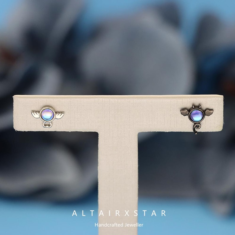 May include: A pair of silver-toned stud earrings. Each earring features a circular iridescent stone with small wing accents. The earrings are displayed on a neutral-colored stand. The text "ALTAIRXSTAR Handcrafted Jeweller" is visible at the bottom.