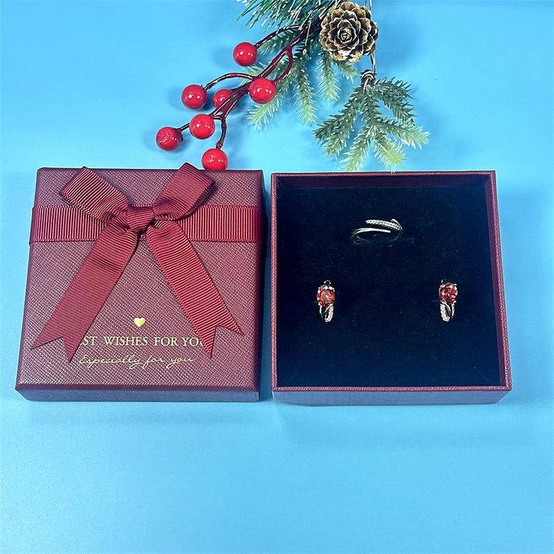 May include: A maroon gift box with a matching ribbon bow, open to reveal a ring and earrings. The earrings and ring have red gemstones. The box has the text "ST. WISHES FOR YOU. Especially for you."