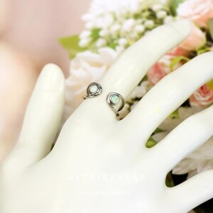 May include: A silver-toned ring with two opal-like stones. The ring has a unique open design, with each stone set in a swirl. The ring is displayed on a white hand, with a soft floral background.