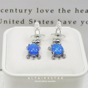 Könnte beinhalten: Ein Paar Ohrstecker mit blauen Opal-Schildkrötenanhängern. Die Ohrringe haben eine silberfarbene Metalleinfassung mit kleinen, klaren Akzentsteinen. Der Text "ALTAIRXSTAR Handcrafted Jeweller" ist sichtbar.