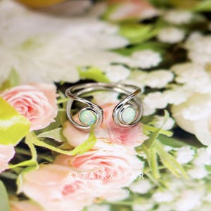 May include: A silver-toned ring with two opal gemstones. The ring has a unique open design, with the gemstones set in circular settings. The ring is displayed on a floral background with pink and white roses.