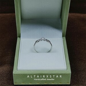 May include: A silver ring with a small, clear gemstone at the center, set in a decorative band. The ring is displayed in a green jewelry box with the text "ALTAIRXSTAR Handcrafted Jeweller" at the bottom.