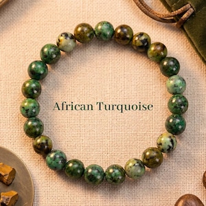 May include: A bracelet made of African turquoise beads. The round beads are polished and display shades of green, brown, and cream. The text "African Turquoise" is below the bracelet.
