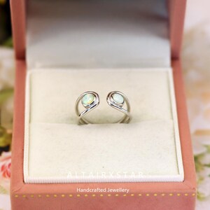 May include: A silver-colored ring with two oval-shaped opal stones. The ring is open at the top and has a unique, swirling design. The ring is displayed in a pink jewelry box. The text "ALTAIRXSTAR Handcrafted Jewellery" is visible.