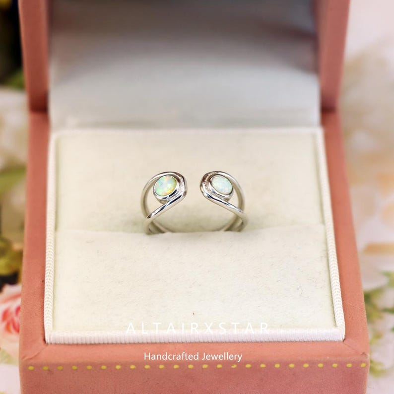 May include: A silver-colored ring with two oval-shaped opal stones. The ring is open at the top and has a unique, swirling design. The ring is displayed in a pink jewelry box. The text "ALTAIRXSTAR Handcrafted Jewellery" is visible.