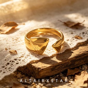 Pode incluir: Um anel aberto de metal dourado polido com duas extremidades triangulares voltadas uma para a outra. A banda tem uma superfície lisa e reflexiva com um espaço entre as duas pontas geométricas. O texto ALTAIRXSTAR está impresso na parte inferior.