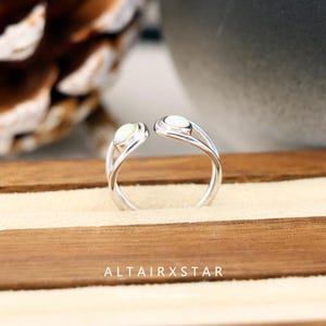 May include: A silver ring with two oval, off-white gemstones. The ring's band curves gracefully, with the gemstones positioned at the open ends. The text "ALTAIRXSTAR" is visible at the bottom of the image. The ring is displayed on a light-colored surface.