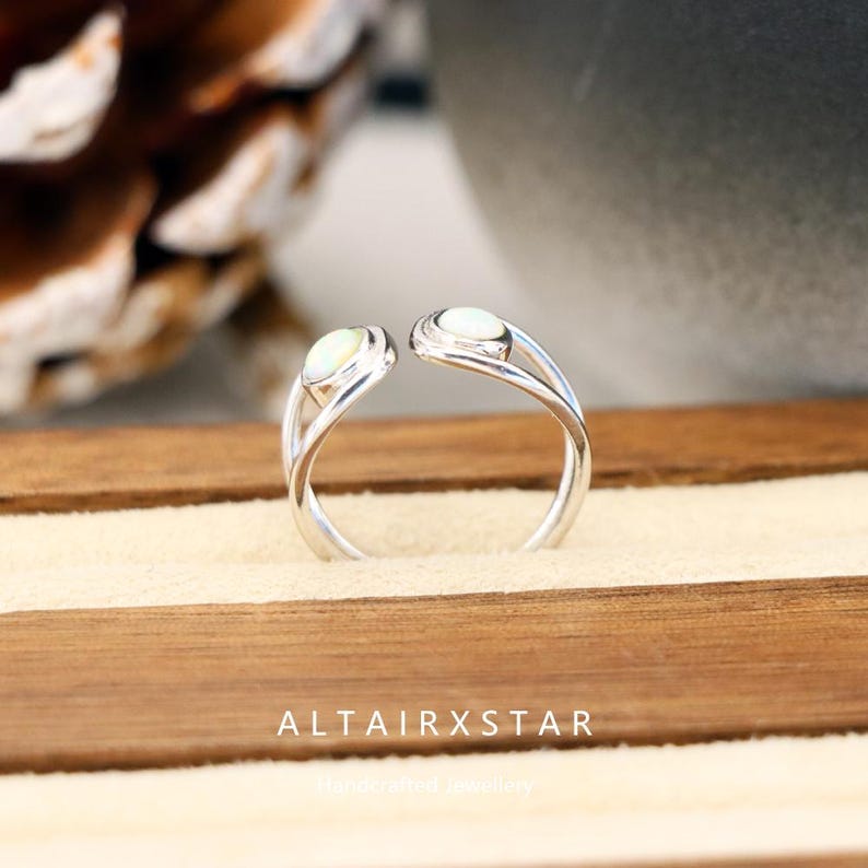 May include: A silver ring with two oval, off-white gemstones. The ring's band curves gracefully, with the gemstones positioned at the open ends. The text "ALTAIRXSTAR" is visible at the bottom of the image. The ring is displayed on a light-colored surface.