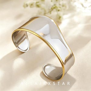 May include: A wide cuff bracelet featuring a polished silver-tone centre with smooth, curved edges and a thin gold-tone border along the top and bottom rims. The open-ended design has a reflective, metallic finish and a continuous, flowing shape.