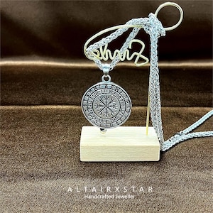 May include: A silver-colored pendant necklace featuring a circular pendant with engraved runes and a central symbol. The necklace is displayed on a wooden block with a gold-colored wire accent. The text "ALTAIRXSTAR Handcrafted Jeweller" is at the bottom.
