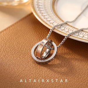 May include: A silver-toned box chain necklace featuring a pendant composed of two interlocking rings. The outer ring is silver-toned, while the inner ring is rose gold-toned and set with a small clear stone. The inner ring is engraved with text.