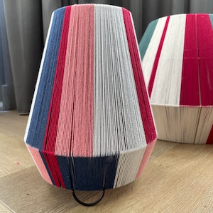 Puede incluir: Dos pantallas de lámpara a rayas hechas de hilo tejido. Las pantallas de lámpara son en tonos de rosa, rojo, blanco, azul y gris. Las pantallas de lámpara están sobre un suelo de madera.