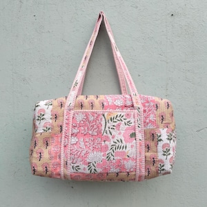 Puede incluir: Bolso tote acolchado con un estampado floral en rosa, blanco y beige. El bolso tiene dos asas y un bolsillo delantero. El bolso es rectangular y parece estar hecho de tela.