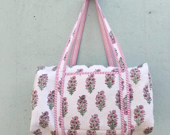 May include: White tote bag with a floral pattern in pink and green. The bag has pink striped handles and trim. The bag is hanging against a light blue wall.