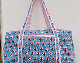 May include: A blue tote bag with a floral pattern in pink and green. The bag has a rectangular shape with a zippered pocket and two handles. The handles and trim are pink and white striped.
