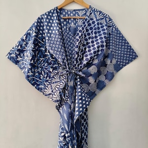 May include: A blue and white patterned kaftan dress with a V-neck and tie closure. The fabric features various designs, including polka dots, swirls, and floral patterns. The dress is hanging on a wooden hanger against a white background.