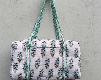 May include: White quilted tote bag with a repeating pattern of blue floral sprigs. The bag has green trim and green striped handles. The bag is rectangular and appears to be made of cotton.
