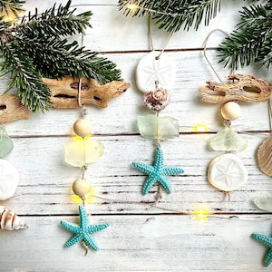 May include: Five handmade Christmas ornaments with a beach theme. Each ornament features a starfish, seashell, sand dollar, and sea glass. The ornaments are strung on twine and are hanging from a white wooden background.