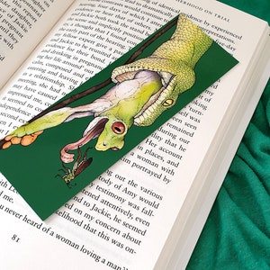 May include: A green bookmark featuring a detailed illustration of a green snake consuming a frog, with a small insect below. The bookmark is placed in an open book with text visible on the pages.