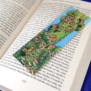 Könnte beinhalten: Ein farbenfroher Lesezeichen mit einer Illustration eines Kaktus mit Vögeln und Blumen. Das Lesezeichen befindet sich in einem Buch. Der Hintergrund ist blau.