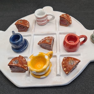 Könnte beinhalten: Ein weißes, teekannenförmiges Keramikbrett mit einem Tic-Tac-Toe-Gitter. Die Spielfiguren bestehen aus Miniatur-Keramikobjekten, darunter verschiedenfarbige Tassen, Becher und braune, dreieckige Kuchenstücke in den Gitterfeldern.