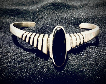 Puede incluir: Una pulsera de plata con una piedra ovalada negra en el centro. La pulsera tiene un diseño geométrico con una serie de líneas en relieve a cada lado de la piedra.