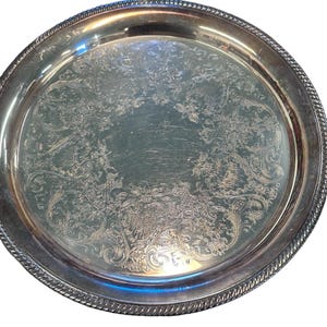 Può includere: Un vassoio da portata in argento con un design decorativo in rilievo. Il vassoio ha un bordo rialzato e un bordo a forma di corda.