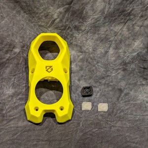 May include: A yellow plastic 3D printed part with two circular openings and a logo on the front. There are three small black and white plastic pieces next to it.