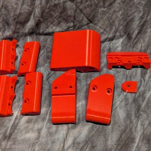 May include: A set of 10 bright red 3D printed plastic pieces. The pieces are various shapes and sizes, including rectangular blocks, curved pieces, and small, round pieces.