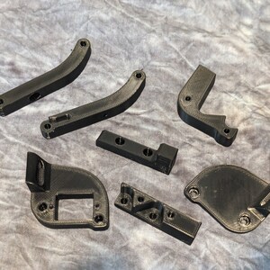 May include: A set of eight black 3D printed plastic parts. The parts are various shapes and sizes, with some having holes and others having raised edges.