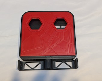 Op de afbeelding: Een rode 3D-geprinte telefoonstandaard met een geometrisch patroon en twee zeshoekige uitsparingen. De standaard heeft een zwarte basis met een standaard.