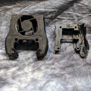 May include: Two black 3D printed parts for a fan mount. The larger part has a fan opening and two arms. The smaller part has a rectangular shape with two arms.