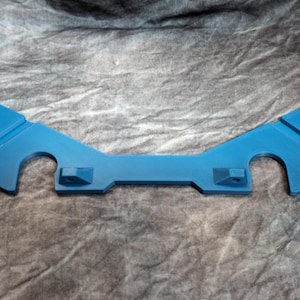 May include: A teal-colored, symmetrical, plastic component with a curved shape and several cutouts. The piece has a central rectangular section with two raised square features and two curved arms extending outwards.
