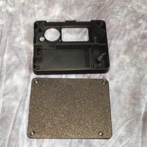 May include: A black rectangular plastic case with a textured, dark gray lid. The case has a circular opening and a rectangular opening. The lid has four screw holes. The case appears to be a protective housing for an electronic device.