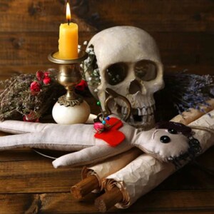 May include: A white human skull with a lit candle in a brass candlestick, a red heart with pins, and two rolled up scrolls on a wooden surface.