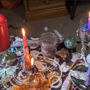 May include: A black tablecloth with a variety of items on it, including candles, a jar of herbs, a crystal, and a piece of sage. The candles are red, purple, and black. The crystal is green. The sage is tied with a white string.