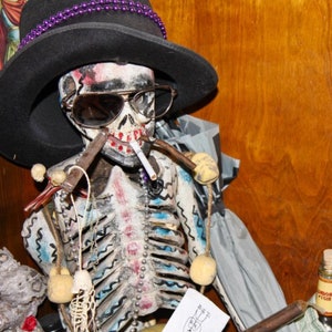 May include: A skeletal figure wearing a black hat with purple beads, sunglasses, and a cigarette. The skeleton is decorated with blue and white paint and has a beaded necklace. The figure is holding a pipe in its right hand.