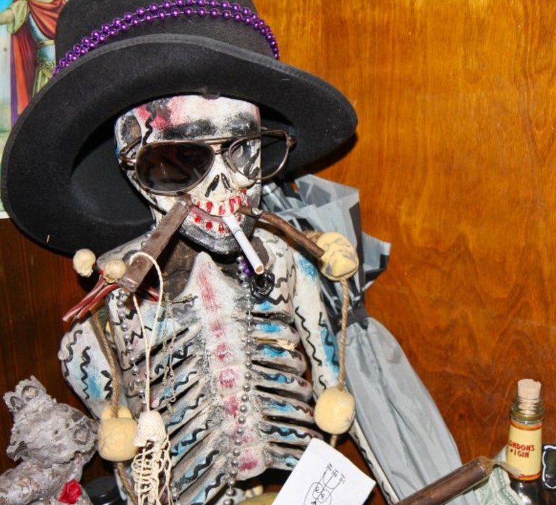 May include: A skeletal figure wearing a black hat with purple beads, sunglasses, and a cigarette. The skeleton is decorated with blue and white paint and has a beaded necklace. The figure is holding a pipe in its right hand.