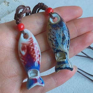 May include: Two ceramic fish pendants with brown cords and red beads. One pendant is white with red and blue patterns, the other is blue and brown with a marbled effect. The pendants have a hole at the top and a small opening at the bottom.