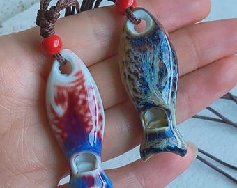 May include: Two ceramic fish pendants with brown cords and red beads. One pendant is white with red and blue patterns, the other is blue and brown with a marbled effect. The pendants have a hole at the top and a small opening at the bottom.