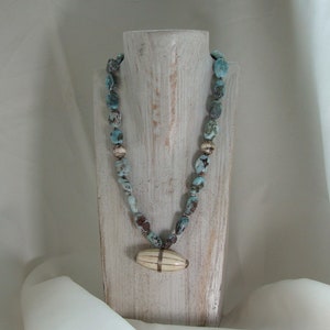 Puede incluir: Un collar de piedras turquesa y marrón con un colgante de hueso blanco. El collar se muestra en un maniquí de madera blanco.