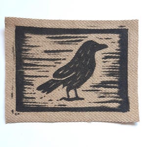 May include: A rectangular fabric patch featuring a black ink block print of a bird in profile. The design is centred on a tan, textured, woven material with a visible diagonal twill pattern. The bird is depicted with simple, bold lines.